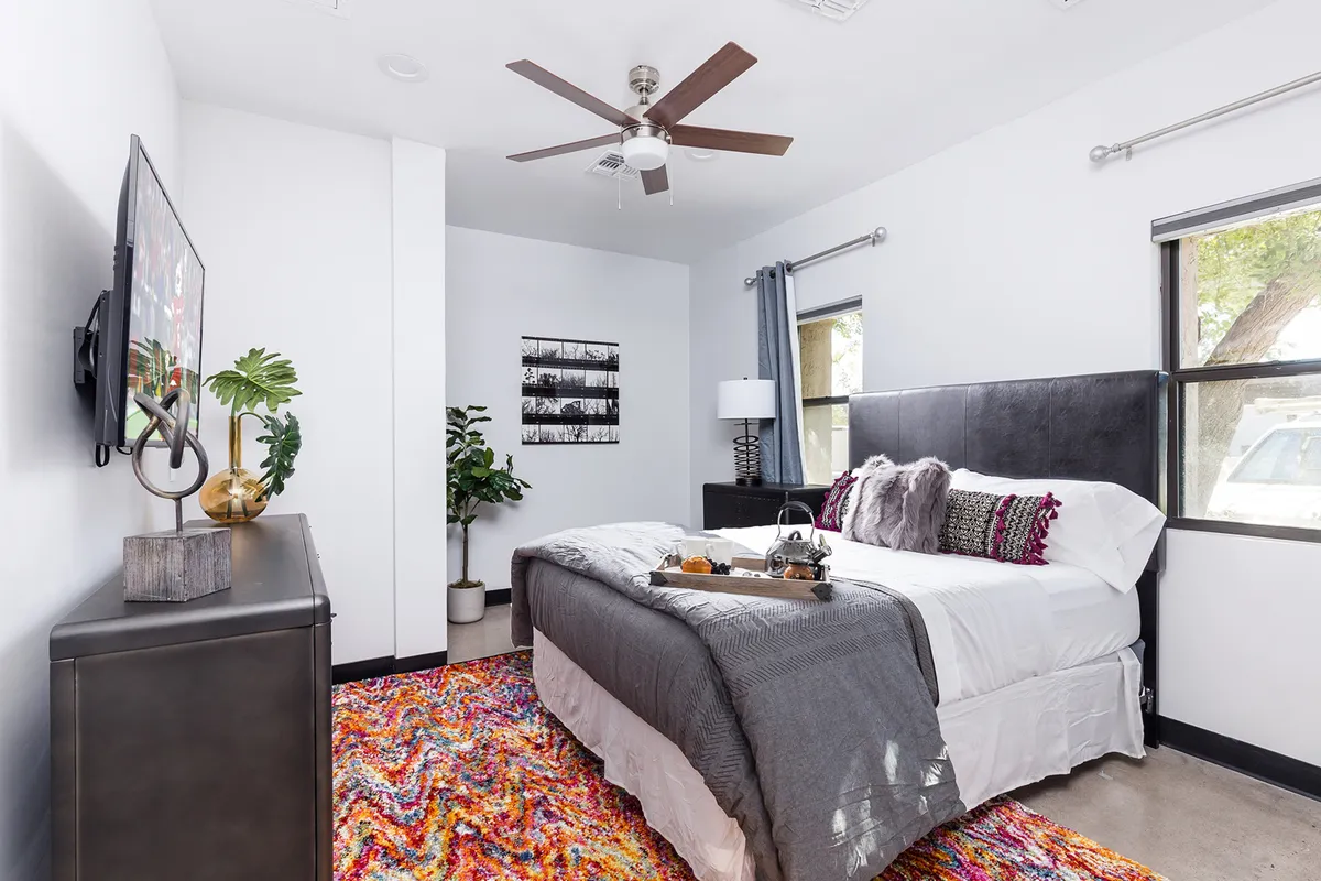 A comfortable bedroom featuring a neatly made bed with decorative pillows, a colorful area rug that adds vibrancy, a wall-mounted TV ideal for unwinding, and a nightstand with a cozy lamp that creates a relaxing atmosphere.