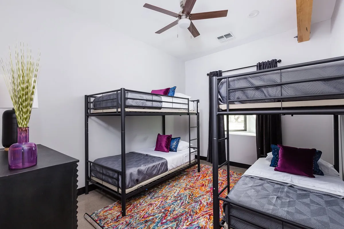 A playful bunk room with two sets of bunk beds, a colorful rug, and decorative pillows, all under a ceiling fan. The room feels energetic and is clearly designed to delight kids and teens.