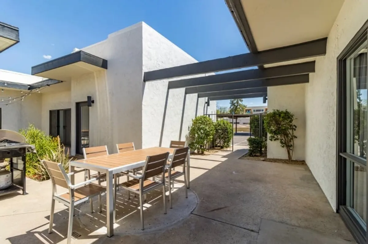 A spacious patio features an outdoor dining table, built-in grill, and shaded seating, surrounded by greenery and distinctive architectural beams. The setup is perfect for relaxed barbecues or lively group celebrations, clearly illustrating the grill amenity and communal gathering space.