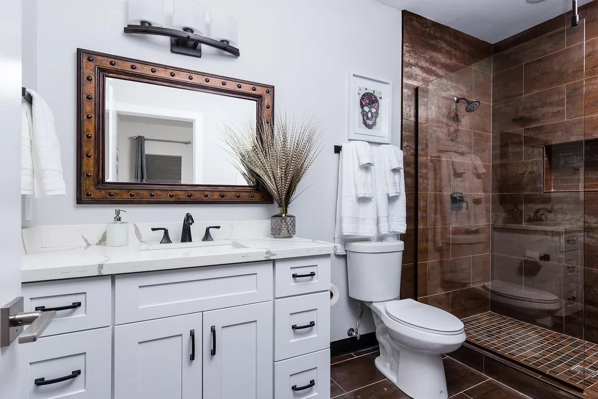 A modern bathroom featuring a large framed mirror, a stylish sink and faucet, an elegant shower area with glass doors, and a tasteful arrangement of decorative plants. The space is bathed in natural light, creating a serene environment.