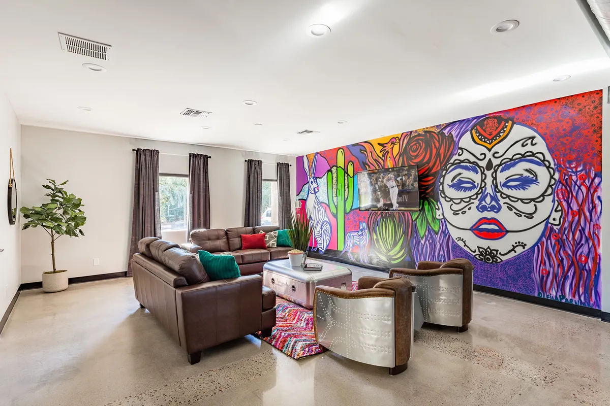 The image showcases a stylish living area featuring a vibrant, colorful mural as the artistic centerpiece, surrounded by plush leather sofas adorned with decorative cushions. The inviting setup is perfect for socializing, watching TV, and relaxing with friends, encapsulating a lively and welcoming ambiance.