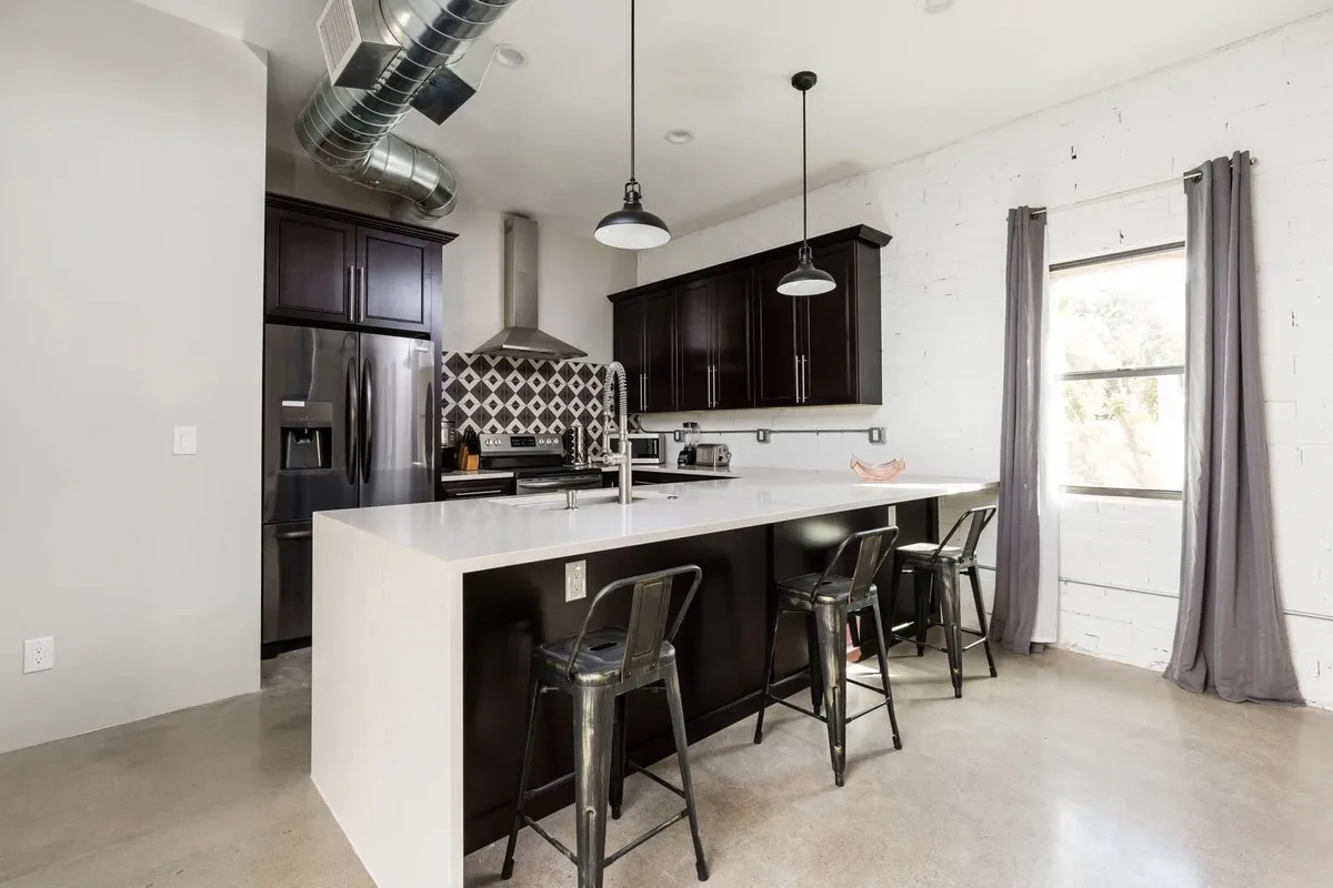 A contemporary kitchen space featuring a prominent kitchen island with black cabinets, a striking geometric backsplash, modern pendant lighting, and bar stools for casual dining. The layout highlights both the ample prep space and the inviting atmosphere for group cooking and entertaining.