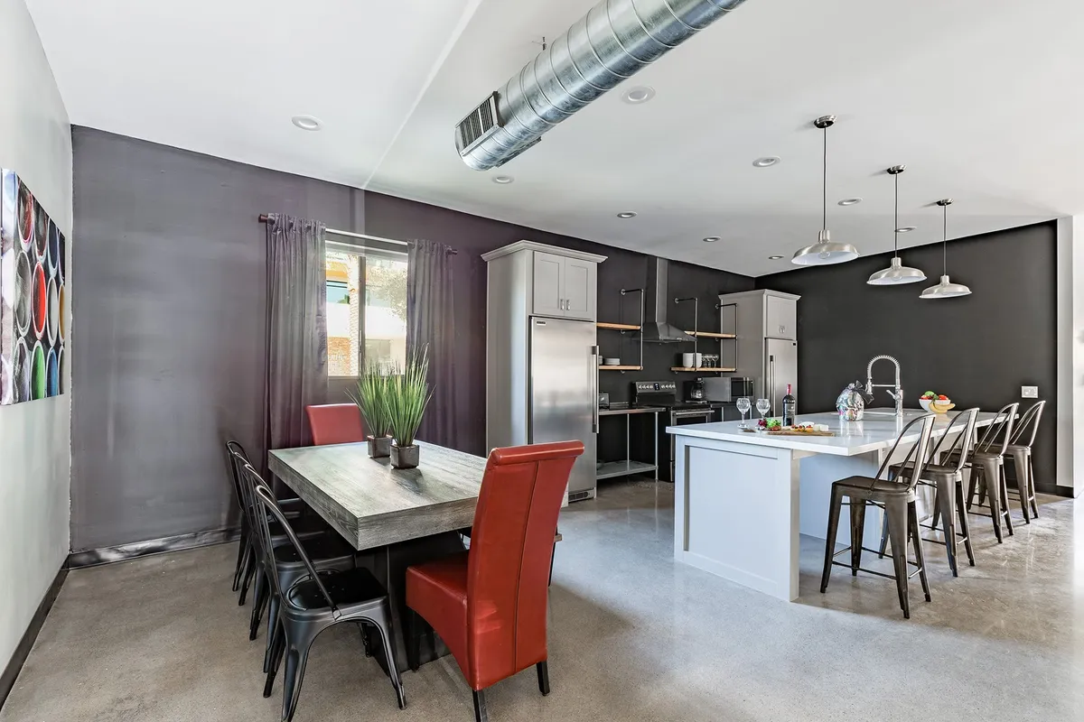 A social dining and kitchen area featuring a large dining table with bold red chairs, a kitchen island with bar stools, and colorful artwork decorating the wall. The open layout encourages communal meals and easy entertaining.